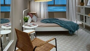 Un dormitorio moderno con cama, una silla, una mesa, una lámpara y vistas al mar a través de grandes ventanales.