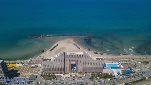 Un hotel grande con una entrada central, un estacionamiento y una zona de playa con sombrillas.