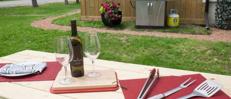 A picnic setup with a bottle of wine, two glasses, a notebook, and barbecue utensils on a wooden table.