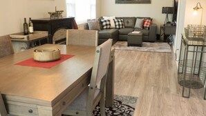 Un coin repas avec une table en bois, des chaises et un set de table rouge. On aperçoit un canapé et un téléviseur à l’arrière-plan.
