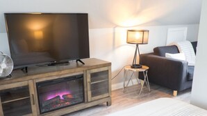 Un salon moderne avec un téléviseur à écran plat posé sur un meuble en bois, une cheminée, une petite table d’appoint avec une lampe et un canapé foncé.