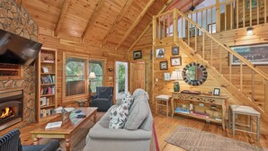 A cozy cabin interior with a fireplace, a sofa, a television, and a staircase. - Dancing Bears | Hot Tub | Grill (Morganton)