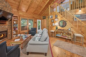 Ein gemütliches Blockhauskaminzimmer mit Kamin, einer Couch, einem Fernseher und einer Treppe.