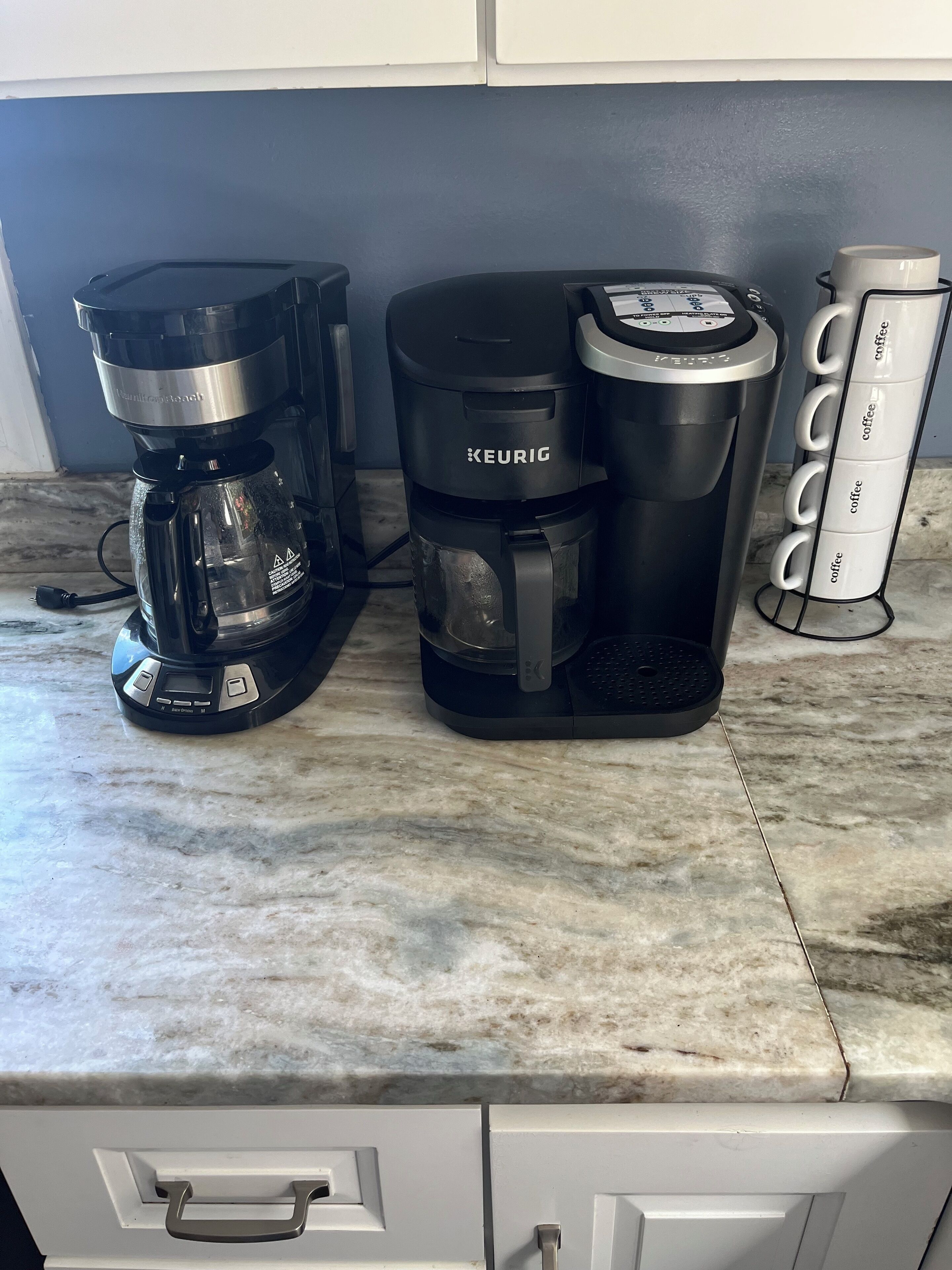 Una cafetera sobre una encimera de mármol, con una máquina de café y un porta-tazas.