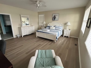 Une chambre à coucher comprenant un lit, des tables de chevet, un ventilateur de plafond, une commode et une fenêtre avec des stores.
