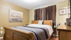 A bedroom with a wooden bed, a nightstand, a desk, a chair, a window with curtains, and a painting on the wall. - Wild North Bed & Rest (Whitehorse)