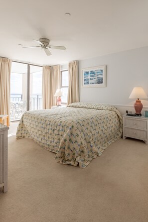 Een slaapkamer met een bed, een plafondventilator, een nachtkastje, een lamp, een ingelijste foto en een balkon met stoelen.