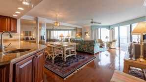 A spacious living room with a large window, a dining area with a table and chairs, and a kitchen with wooden cabinets and a sink. - Surfmaster - Oceanfront 3 Bedroom/3 Bath  Beautiful!!! Great Rates!! (Garden City)