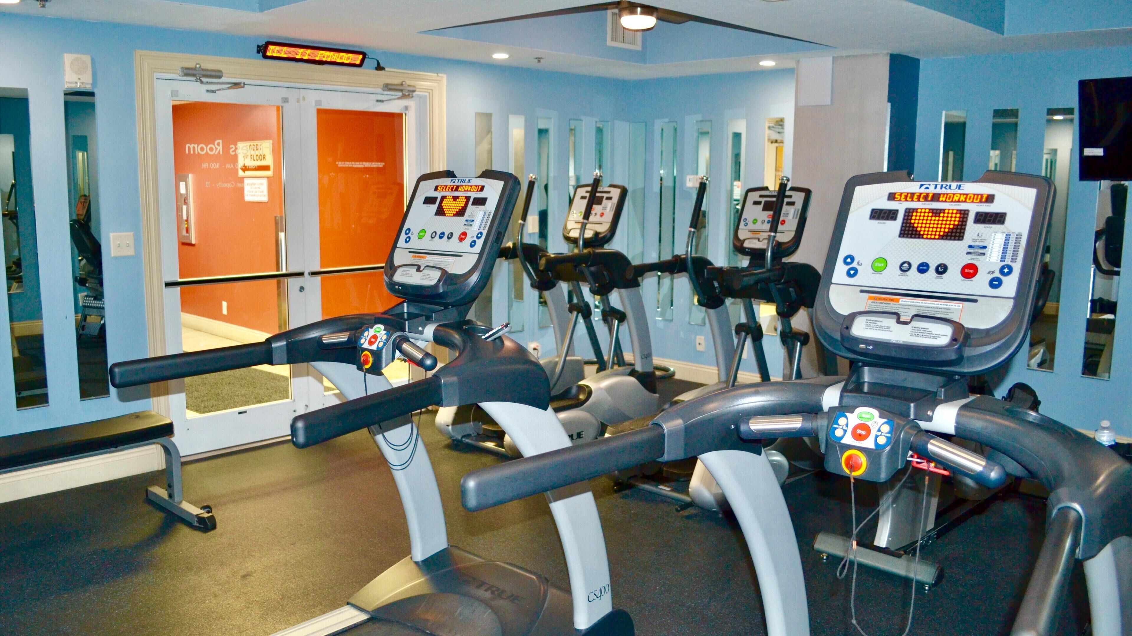 Un gimnasio con cintas de correr y bicicletas estáticas, un muro de espejos y una puerta que indica "1er piso".