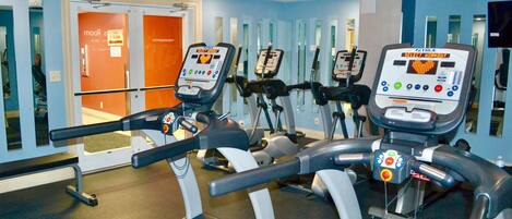 Un gimnasio con cintas de correr y bicicletas estáticas, un muro de espejos y una puerta que indica "1er piso".