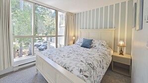 A bedroom with a large window, a bed with floral bedding, a nightstand with a lamp, and a striped wall. - Oceanfront 2 bedroom Condo w/Stunning Views~Heated Pool/Hot Tub/Dock (Nanaimo)