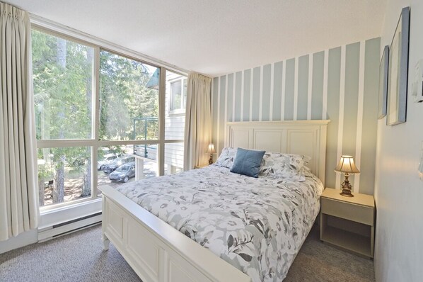 A bedroom with a large window, a bed with floral bedding, a nightstand with a lamp, and a striped wall.