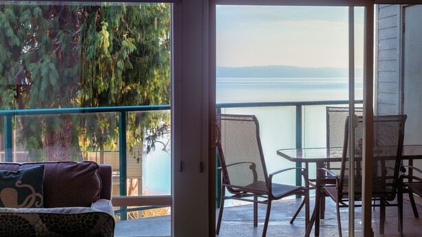 A room with a sliding glass door leading to an outdoor patio with a view of a lake and mountains. - Oceanfront 2 bedroom Condo w/Stunning Views~Heated Pool/Hot Tub/Dock (Nanaimo)