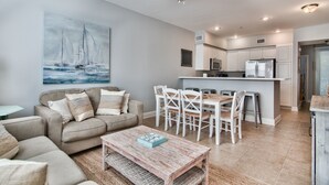 Een moderne woonkamer met een bank, salontafel en een eethoek. Achtergrond toont een keuken met witte kasten en apparatuur.