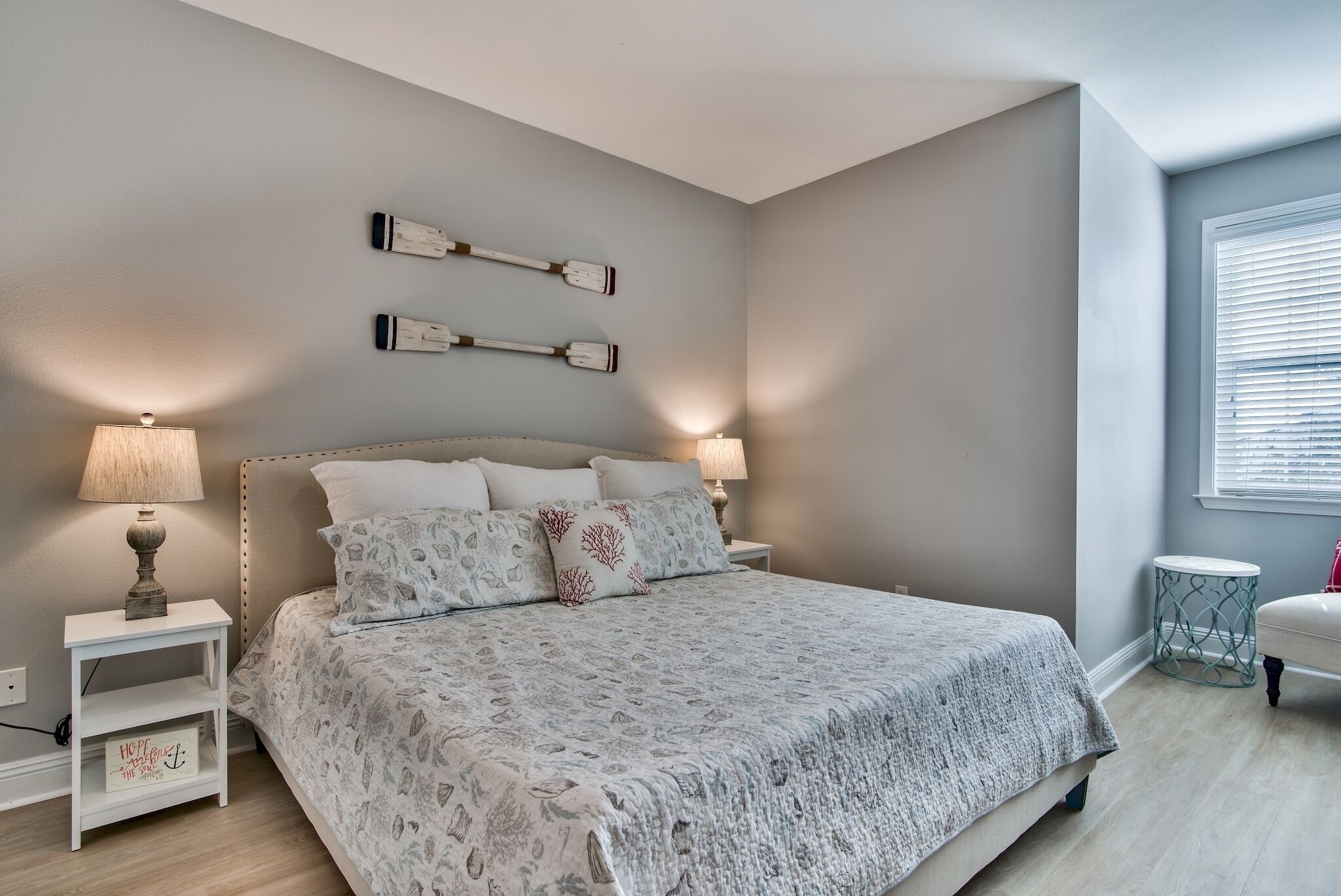 Une chambre à coucher avec un grand lit, deux tables de chevet avec des lampes, une petite table d’appoint et une fenêtre avec des stores.
