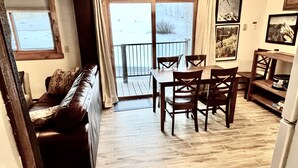 A cozy living area with a brown leather sofa, wooden dining table, and chairs. A sliding glass door leads to a balcony with a view of a snowy landscape. - 2 Bedroom 2 Bathroom Condo - Base Area - Three Seasons (Mount Crested Butte)