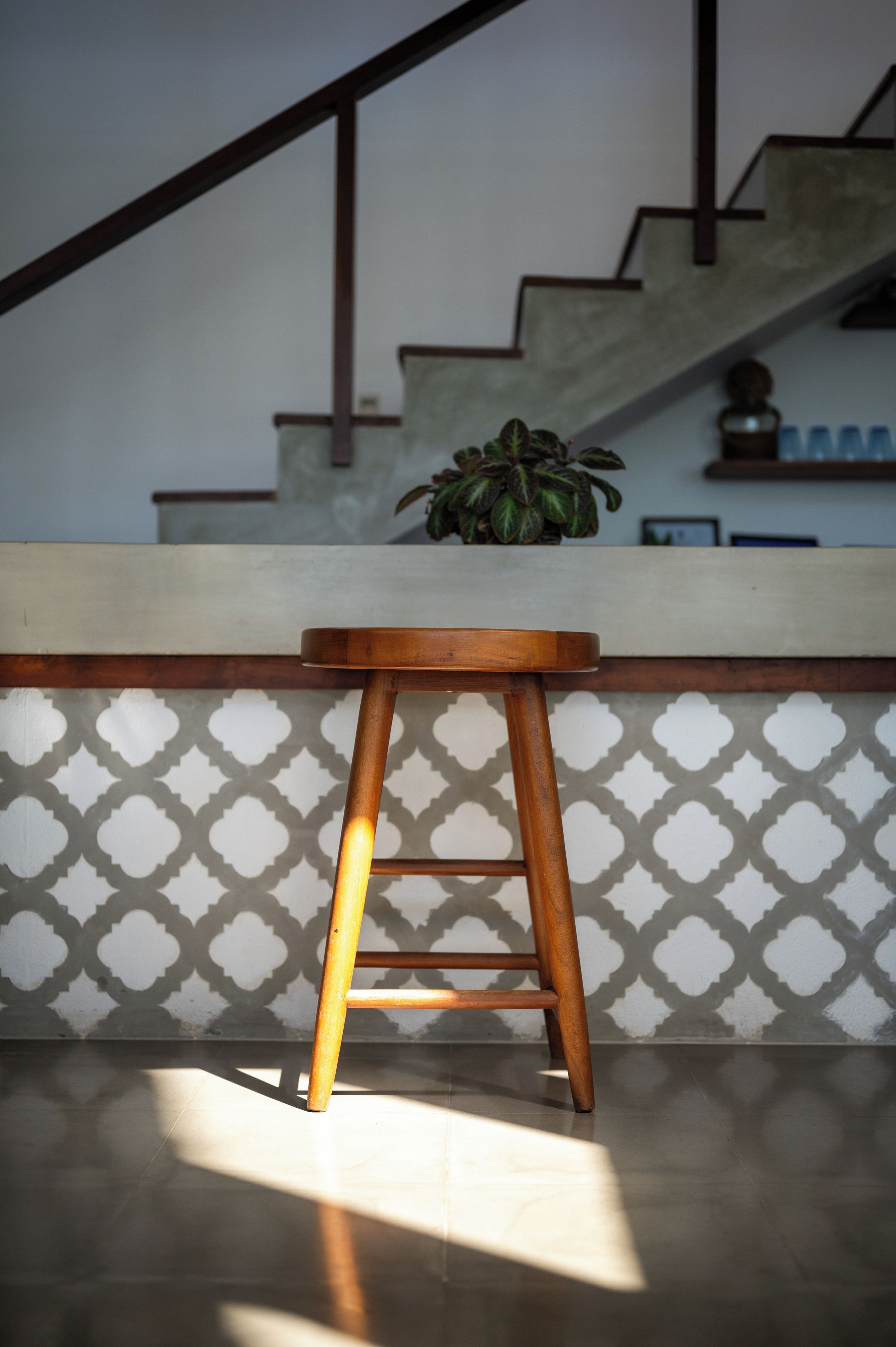 A wooden stool in a room with a patterned wall and a staircase in the background.