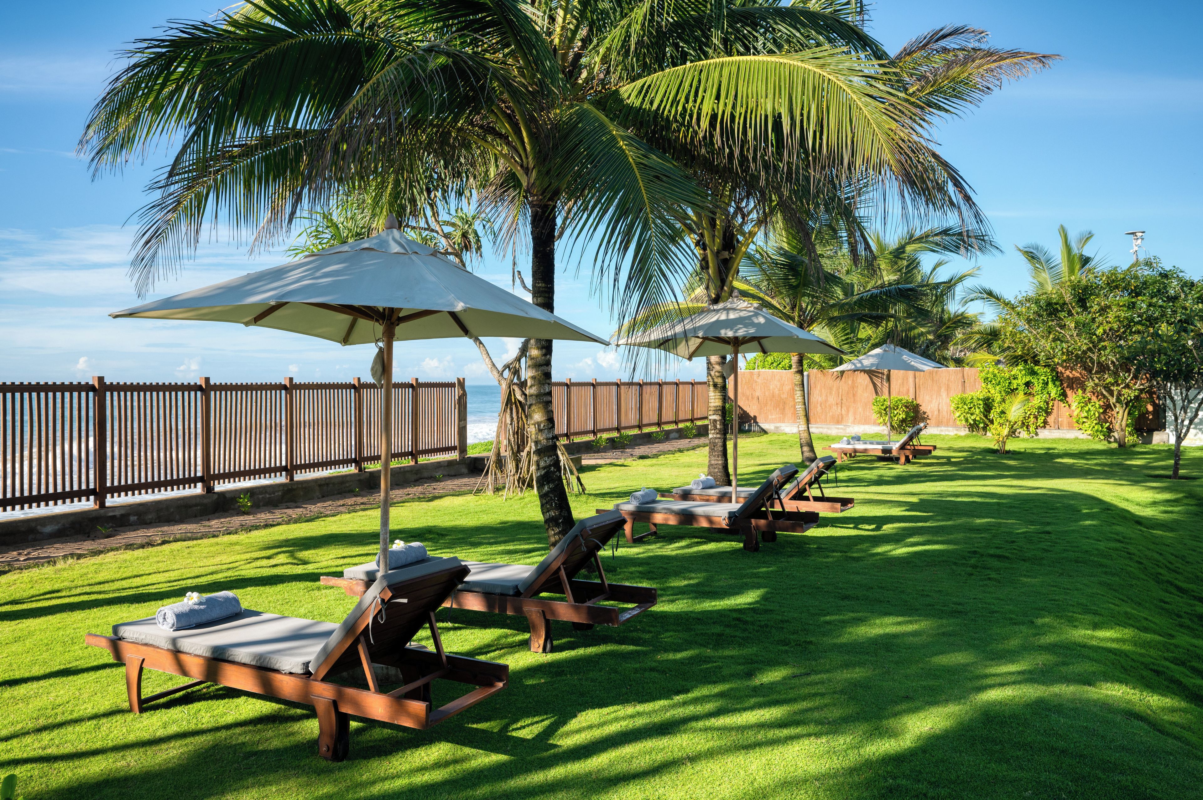 Un área junto a la piscina con sillones de descanso y sombrillas, una valla de madera y palmeras.