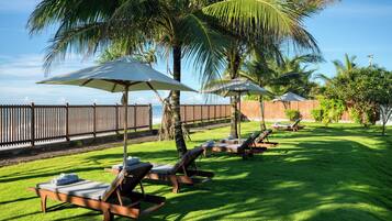Un área junto a la piscina con sillones de descanso y sombrillas, una valla de madera y palmeras.