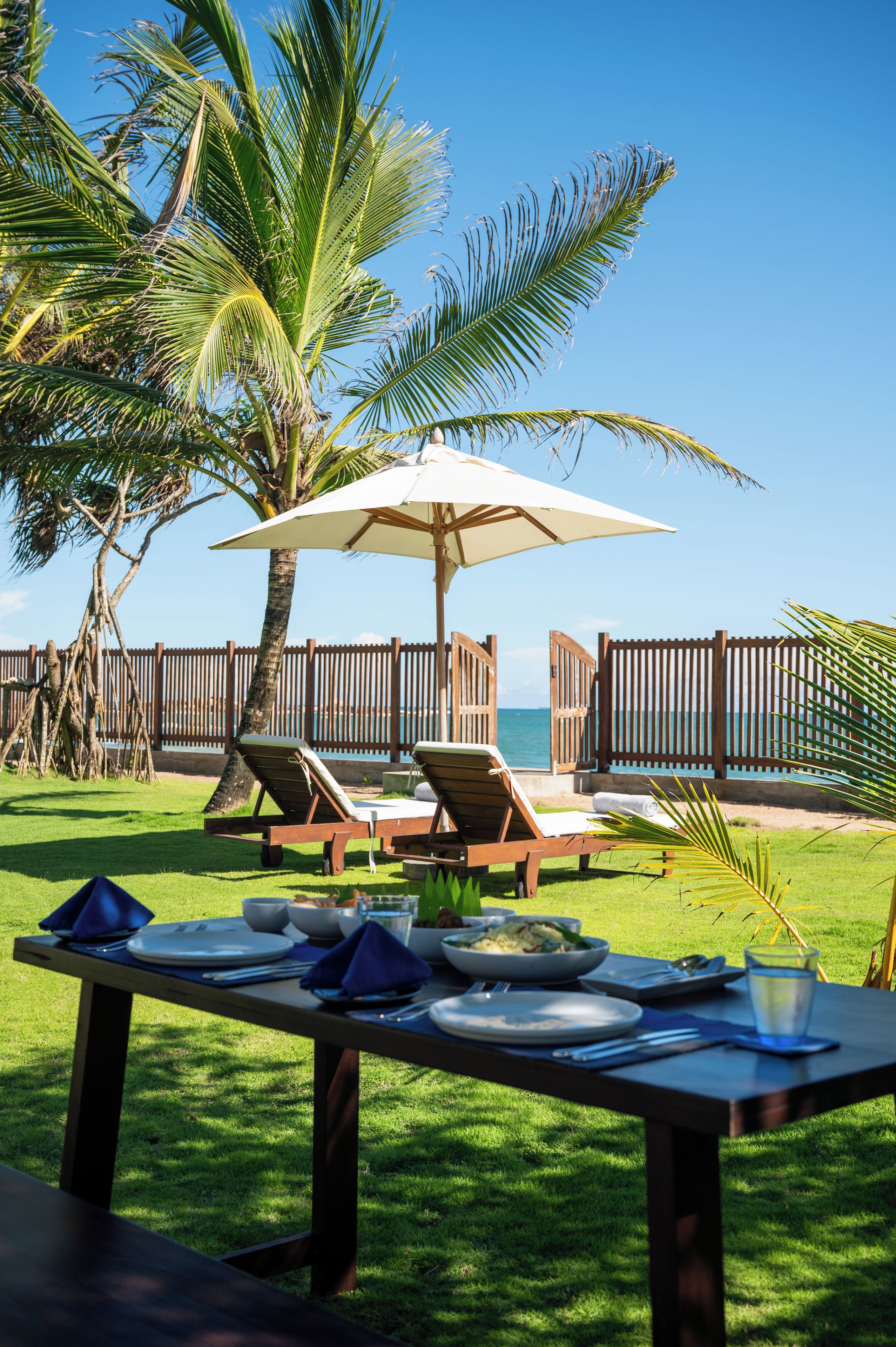 An outdoor dining area with wooden tables and chairs, a large umbrella, and a view of the ocean.