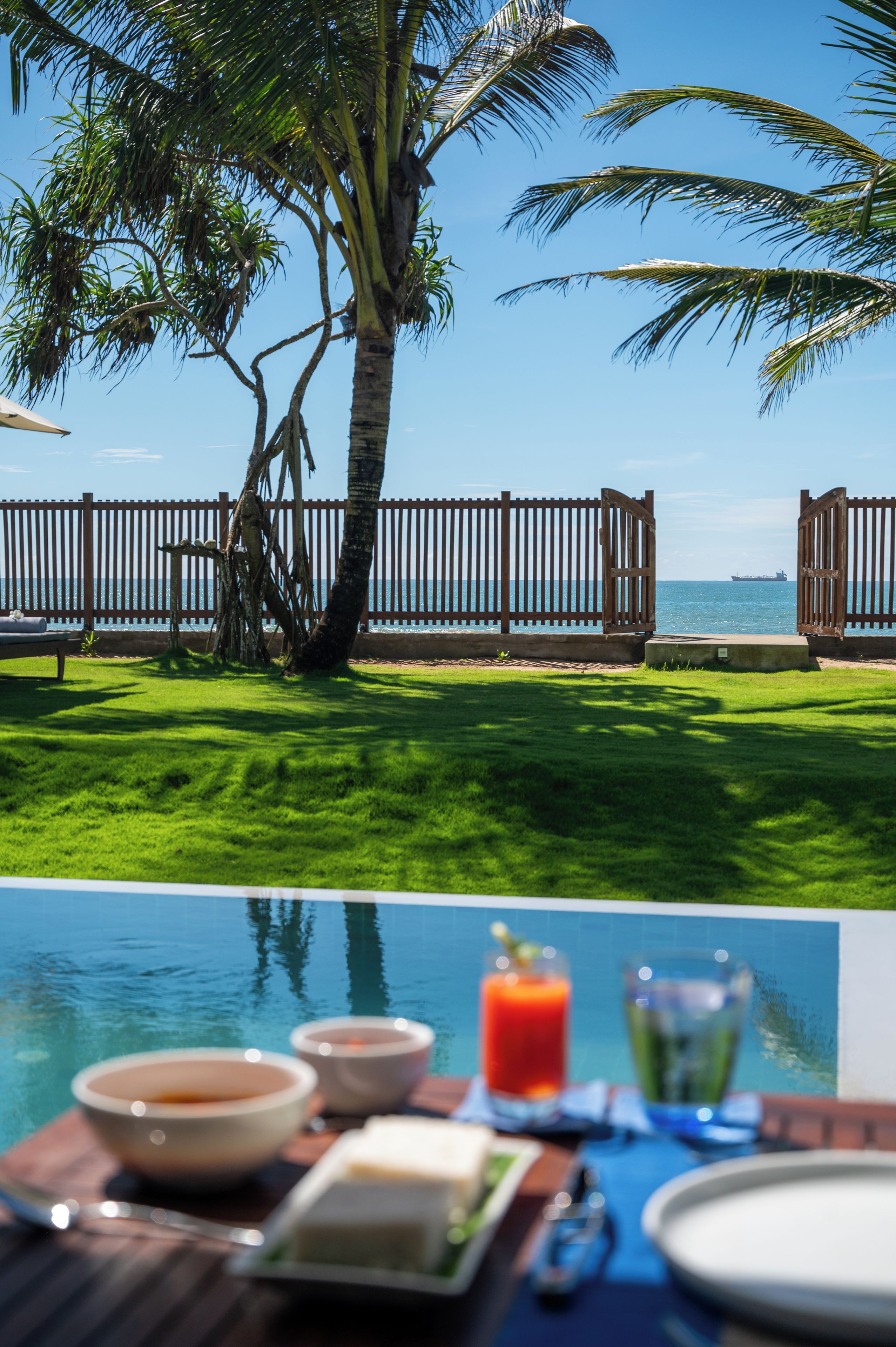 A poolside view with a wooden fence, palm trees, and a clear view of the ocean.