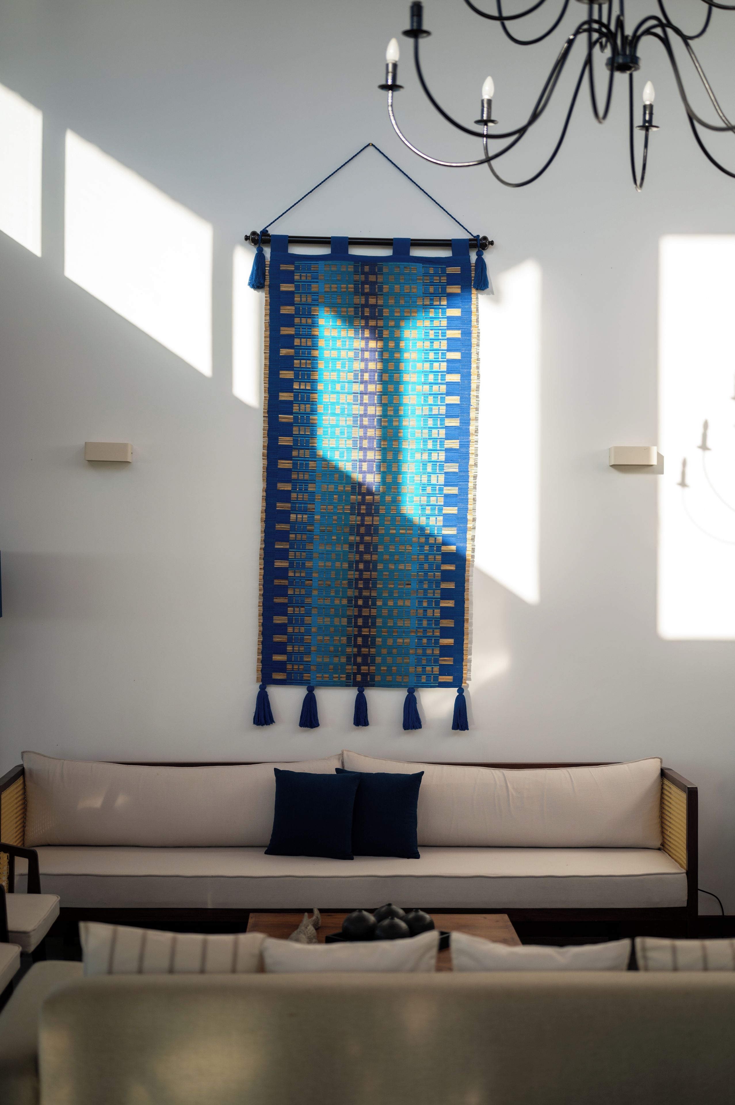 A room with a wall-mounted tapestry, a sofa with cushions, and a chandelier.