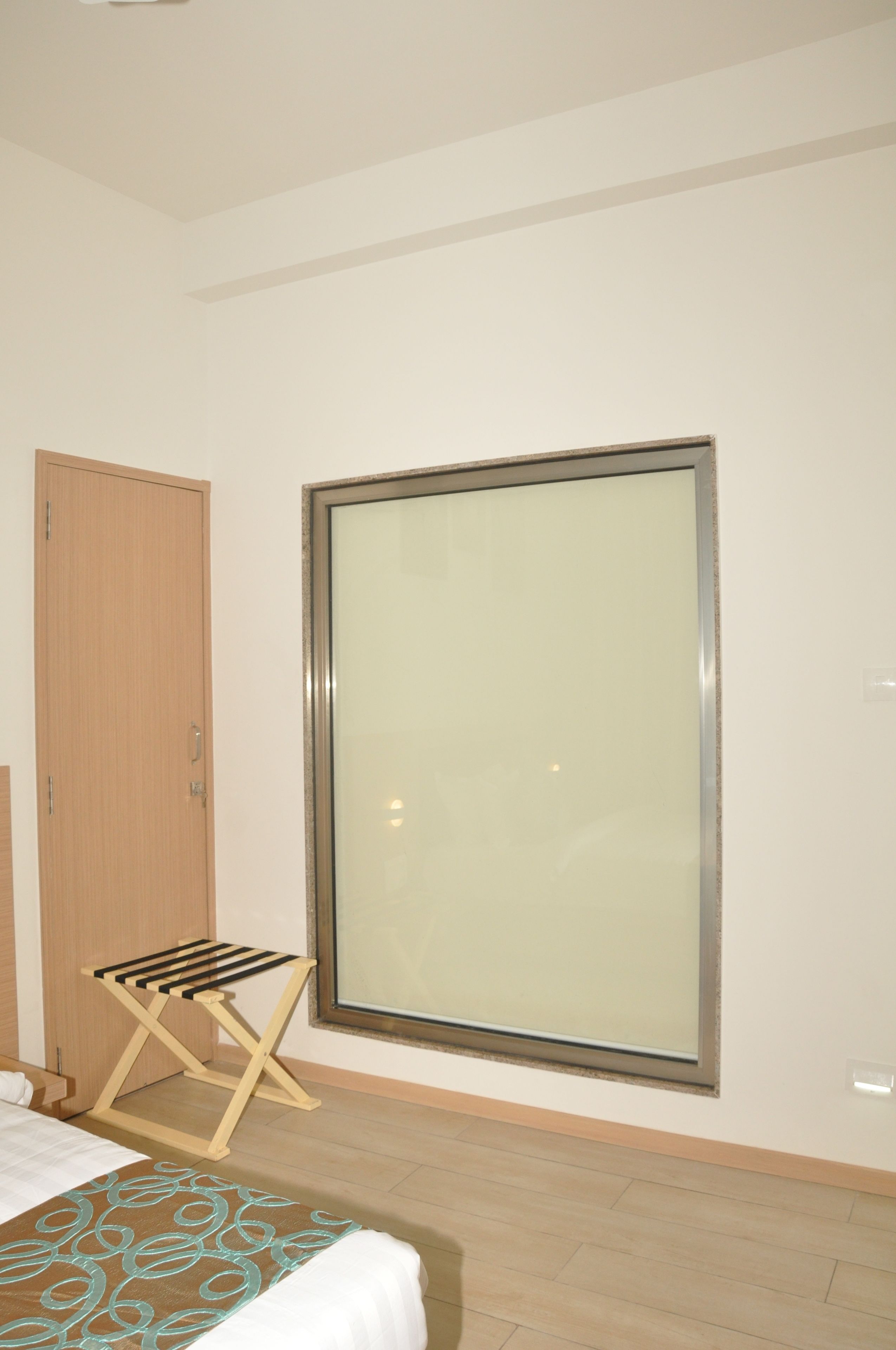 A hotel room with a bed, a wooden wardrobe, a window with a view, and a small wooden stool.