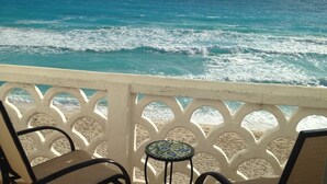 Un balcón con dos sillas y una mesita pequeña, con vista a una playa de aguas turquesas y olas.