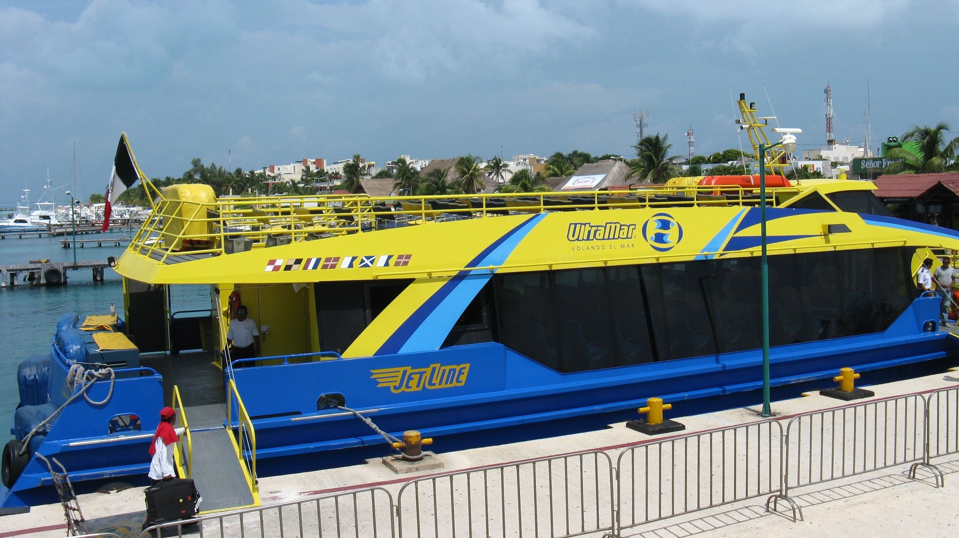 A large blue and yellow ferry with 'Ultramar' branding, docked at a marina with other boats and a clear sky.