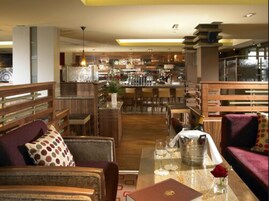 Un moderno vestíbulo de hotel con zona de bar, muebles de madera y un separador de vidrio.