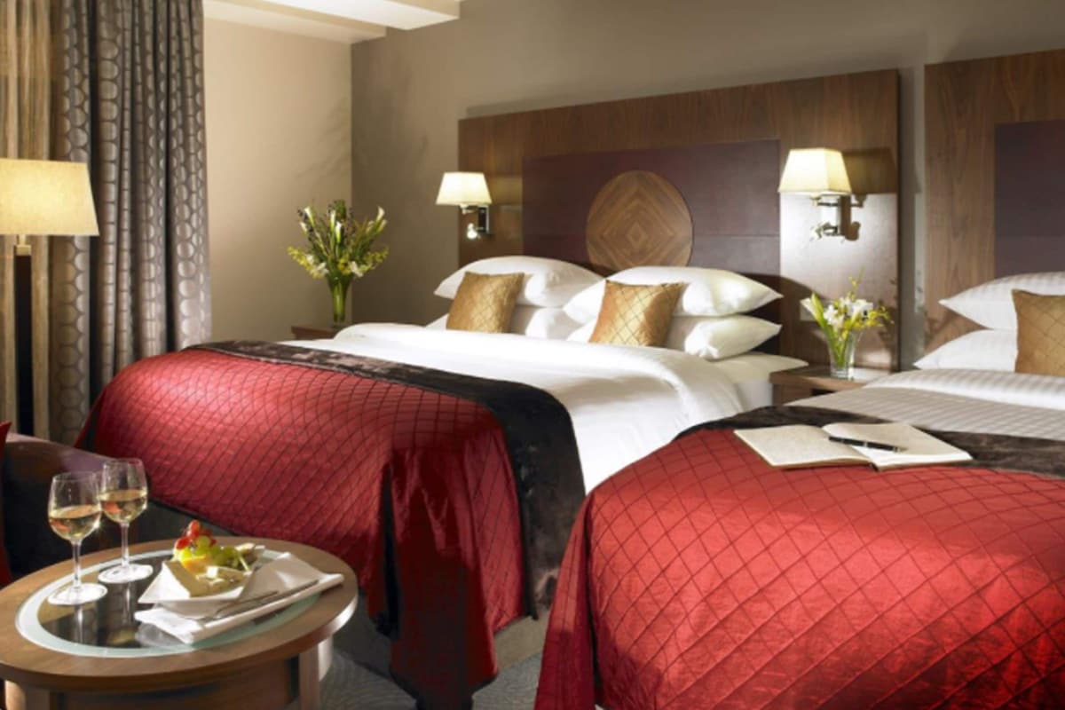 Ein Hotelzimmer mit zwei Betten, einem kleinen Tisch mit einer Tafel mit Speisen und Getränken sowie je einer Lampe auf den Nachttischen.