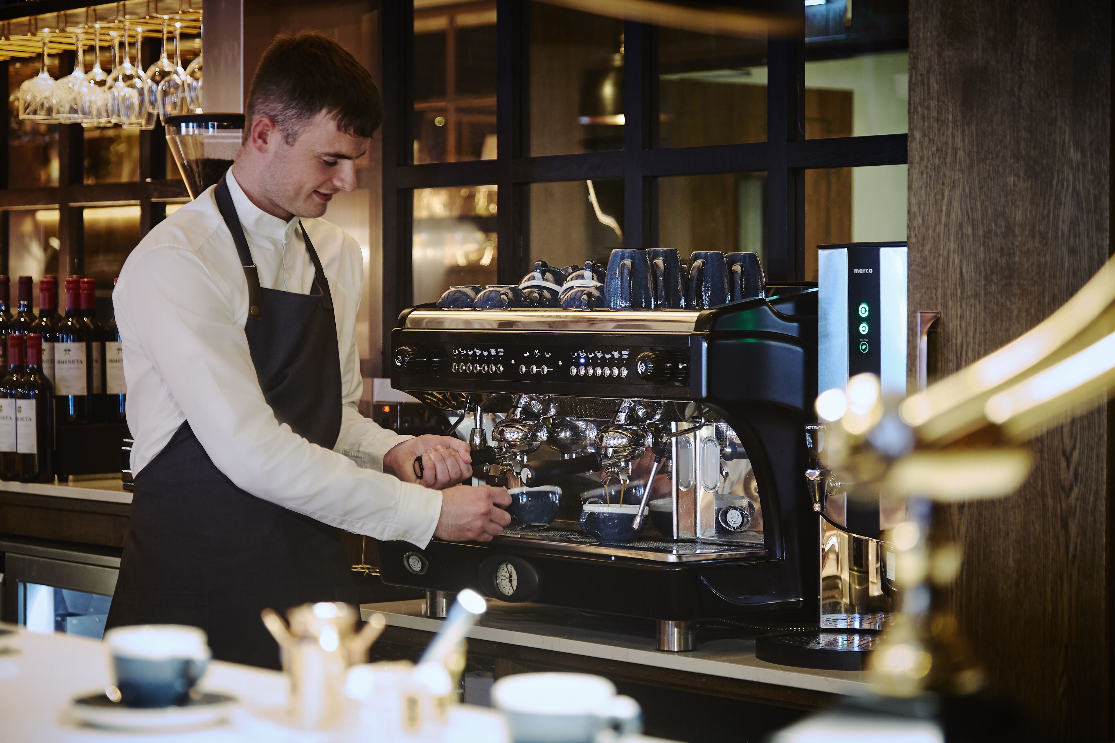 Un barista preparando café en una máquina de espresso comercial con pantalla digital, en un ambiente interior bien iluminado con una vista de botellas de vino al fondo.