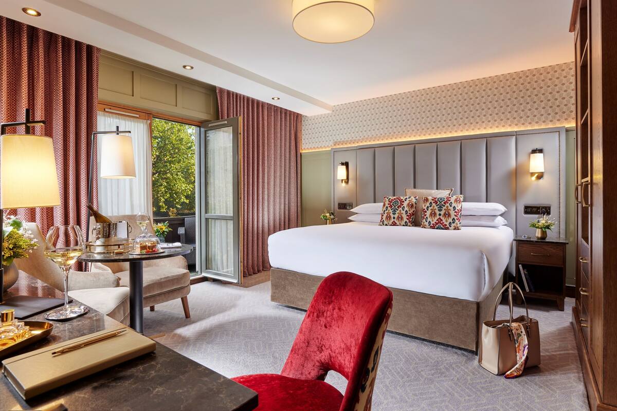 Ein Hotelzimmer mit einem großen Bett, einem Schreibtisch mit Lampe, einem Sessel und Blick auf Grünfläche.