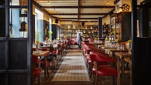 Un restaurant avec des tables en bois et des chaises rouges, un sol à motifs et un coin bar avec des étagères remplies de bouteilles et de verres.