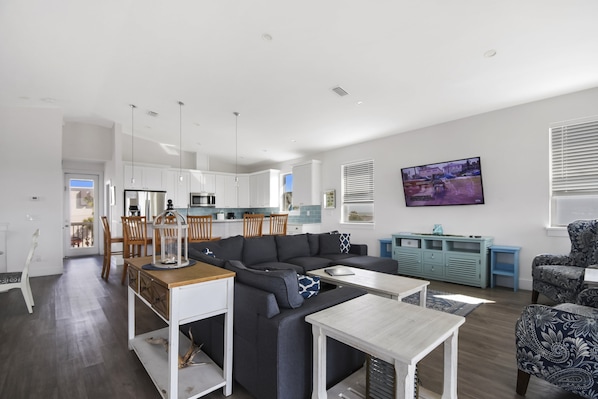 Een moderne woonkamer met een modulaire bank, een eethoek met houten stoelen, een keuken met witte kastjes en een aan de muur bevestigde televisie.