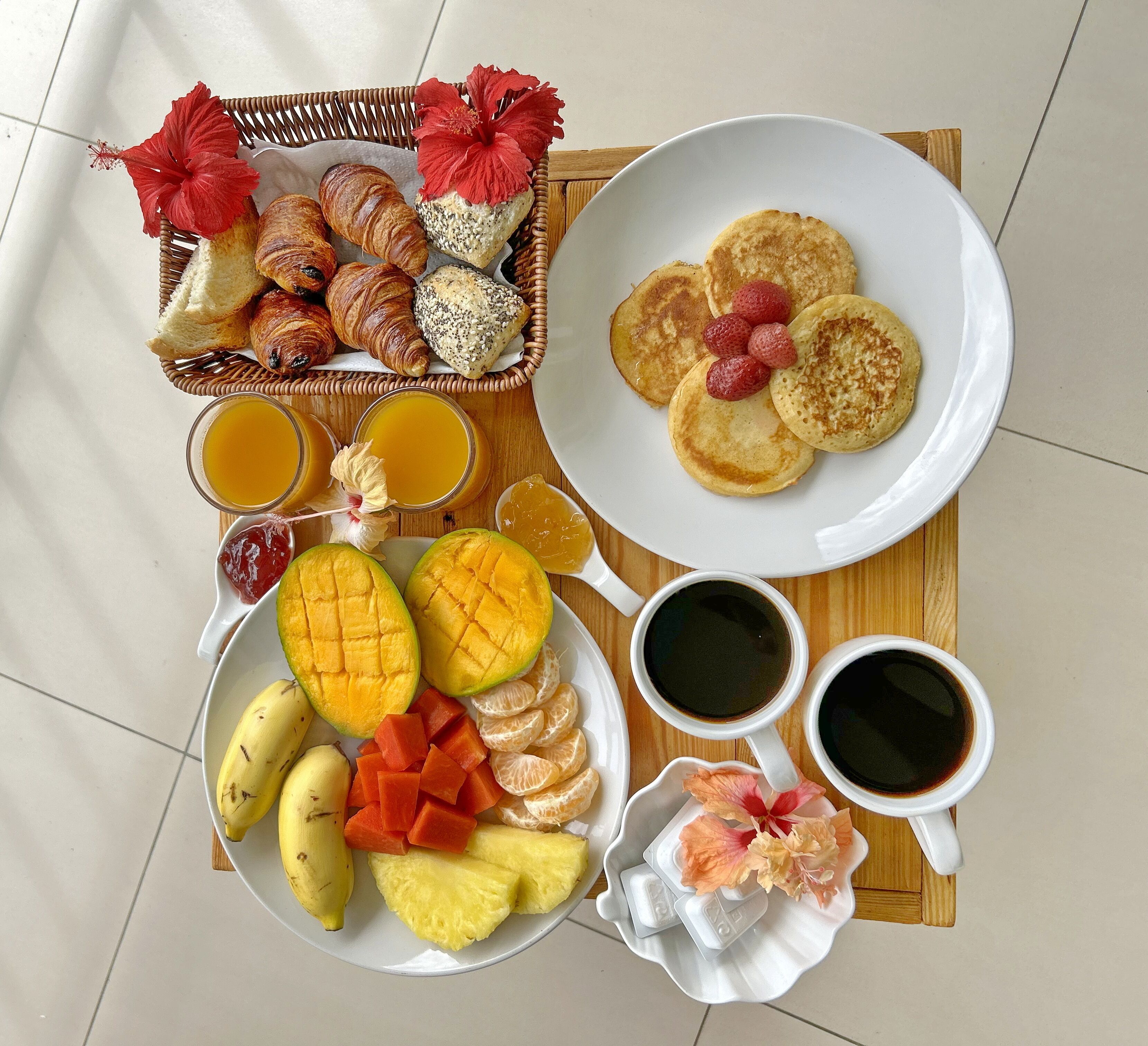 Ein reichhaltiges Frühstücksbuffet mit Croissants, Pfannkuchen, frischem Obst und Getränken auf einer Holztischplatte.