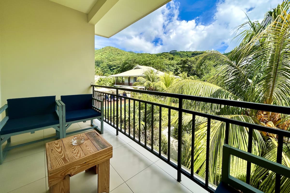 Un balcon avec deux chaises et une petite table, donnant sur un paysage verdoyant et luxuriant, sous un ciel dégagé.