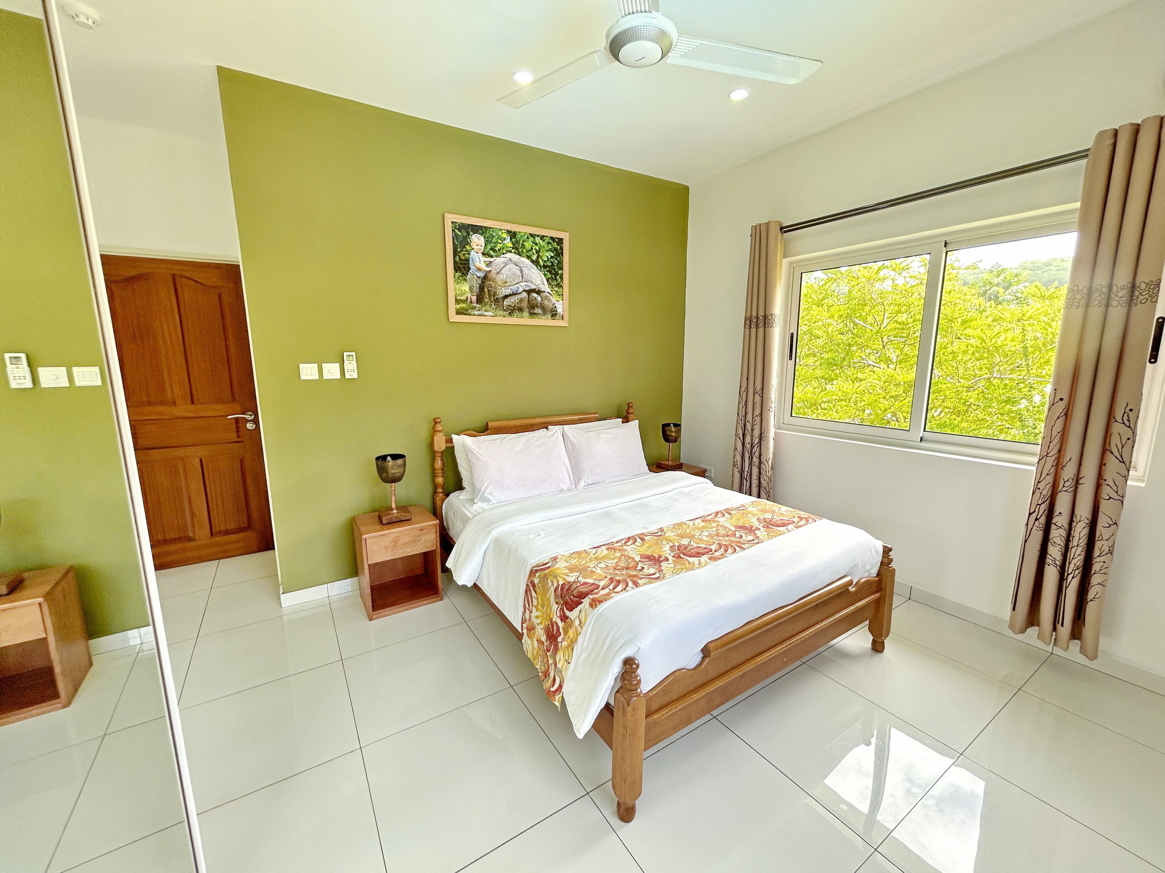 Quarto com cama de madeira, mesa de cabeceira, luminária na parede, quadro emoldurado e janela com cortinas.