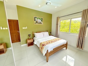 A bedroom with a wooden bed, a nightstand, a wall-mounted lamp, a framed picture, and a window with curtains.
