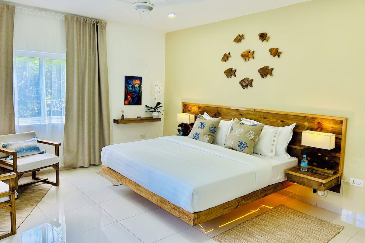 Une chambre d’hôtel moderne avec un grand lit, une tête de lit en bois, une table de chevet avec une lampe, une chaise, une fenêtre avec des rideaux et un mur décoré de motifs de poissons.