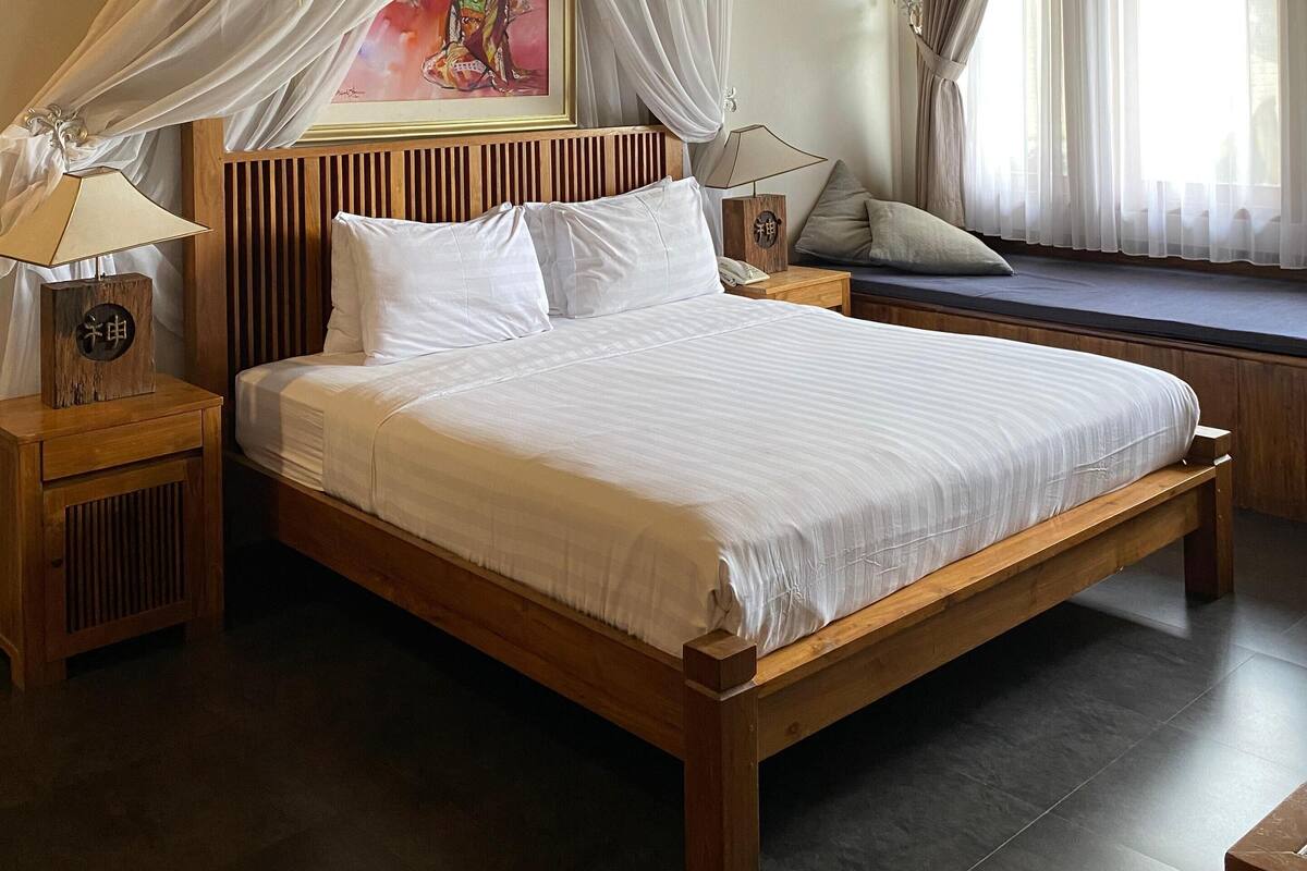 A bedroom with a wooden bed, white bedding, a nightstand with a lamp, a window with curtains, and a painting on the wall.