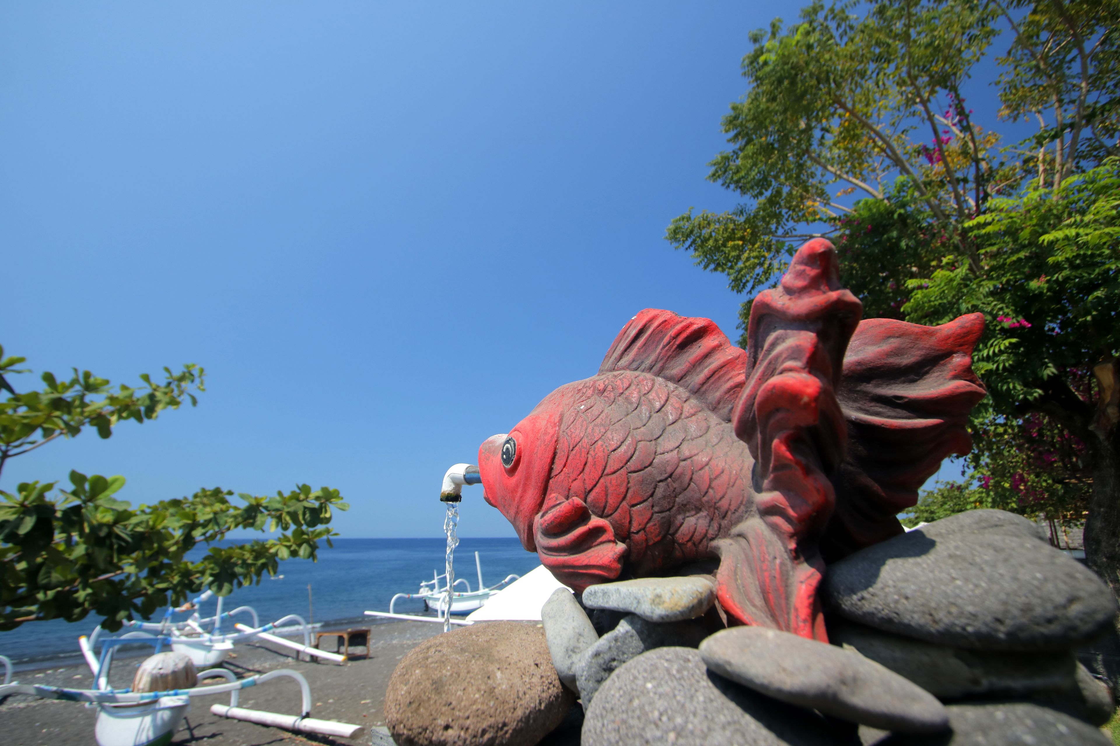 A large red fish sculpture on a rocky shore with boats in the background.