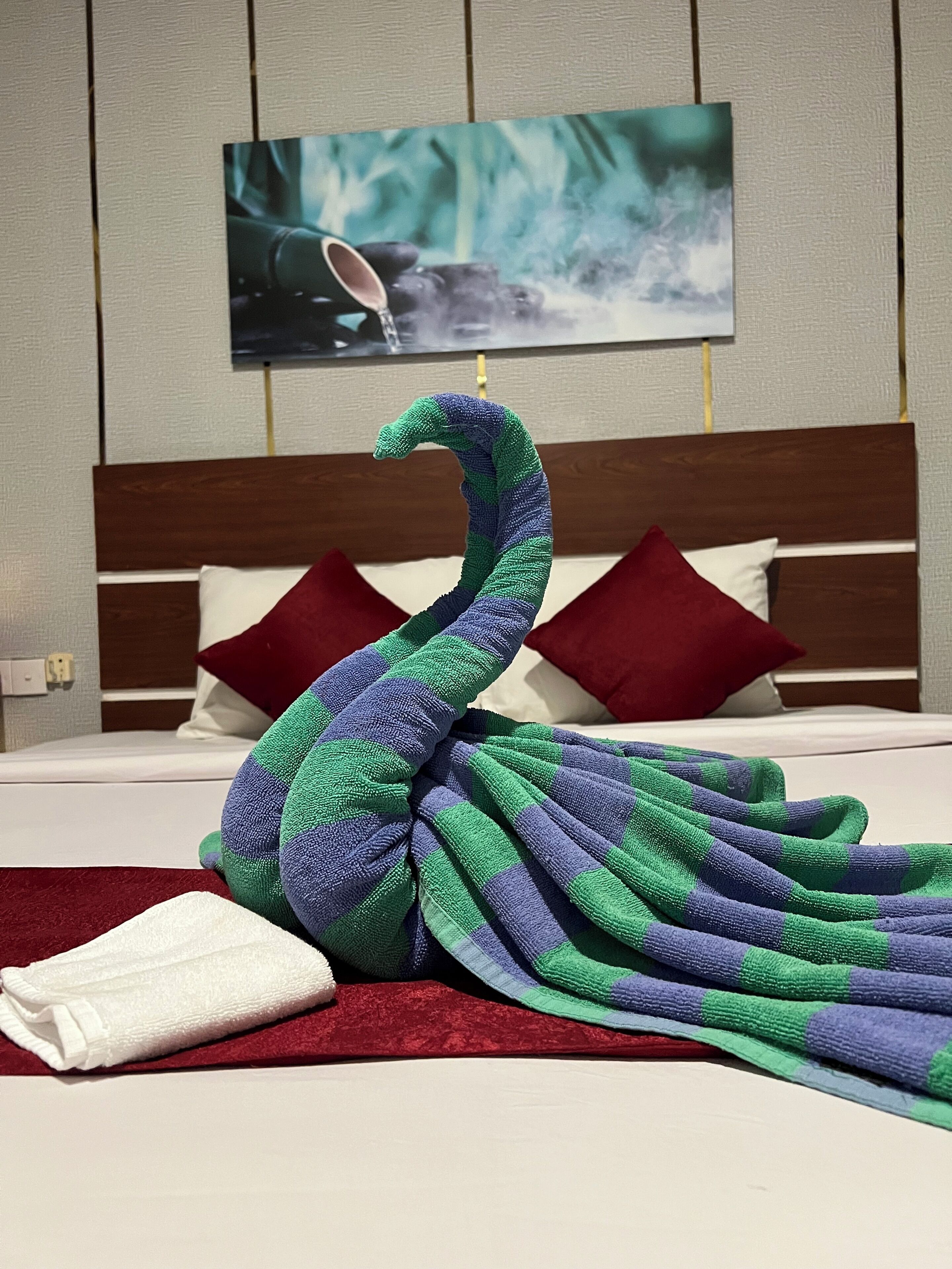 A bed with a peacock-shaped towel, a red pillow, and a white pillow.