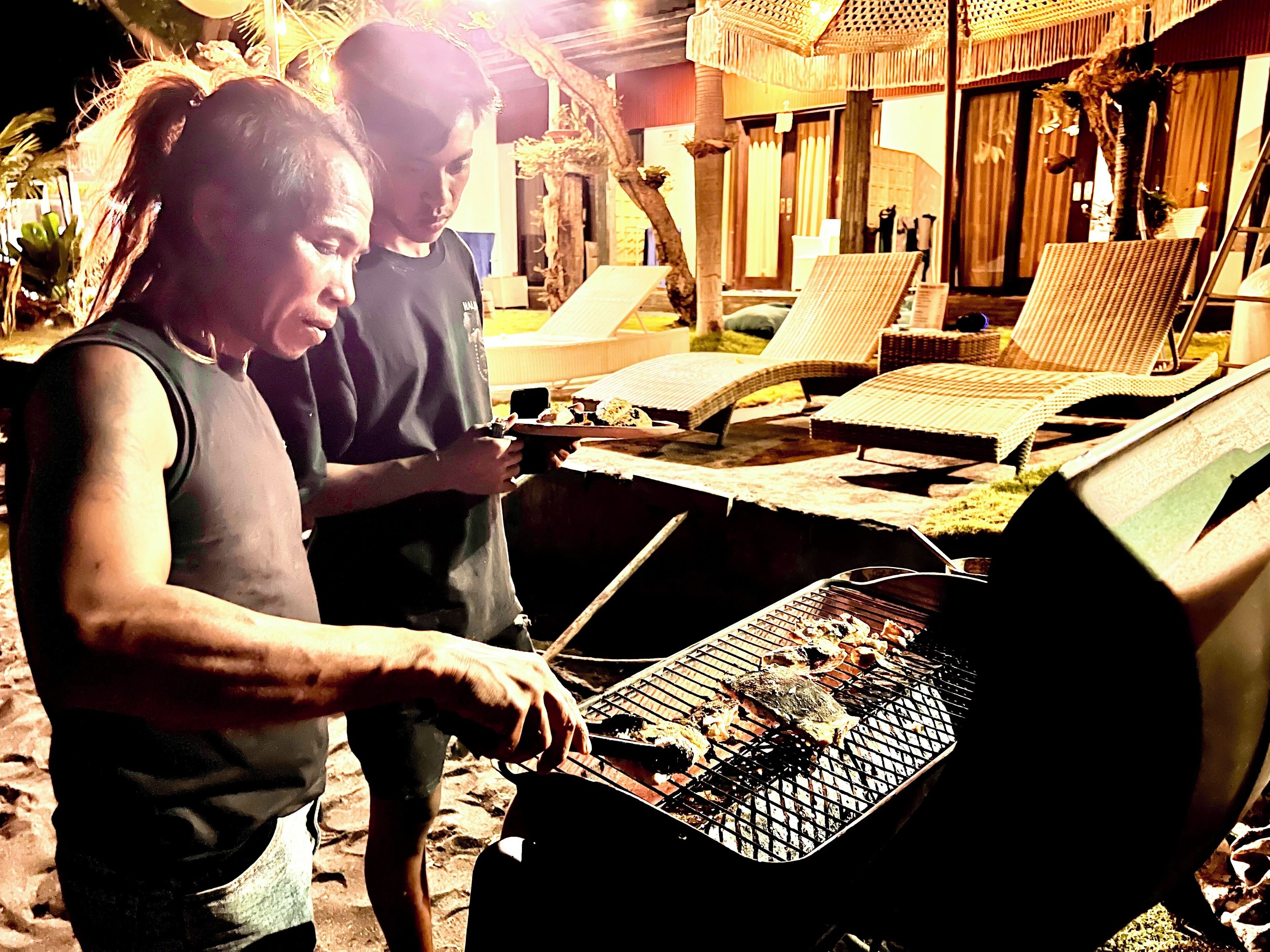 Two people grilling outdoors with lounge chairs and a tiki hut in the background.