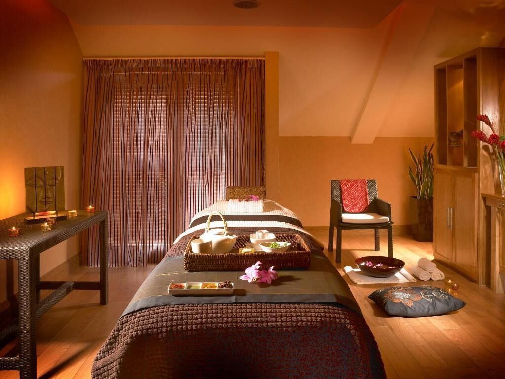 Un cuarto de spa con una camilla de masaje, velas, una silla y una mesita con toallas y un bol.