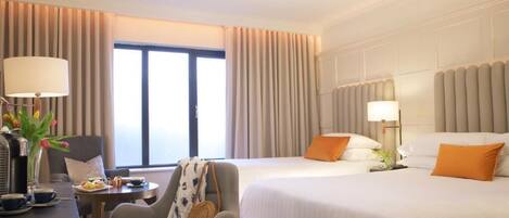 Habitación de hotel con dos camas, un escritorio con lámpara, una silla y un ventanal con cortinas.