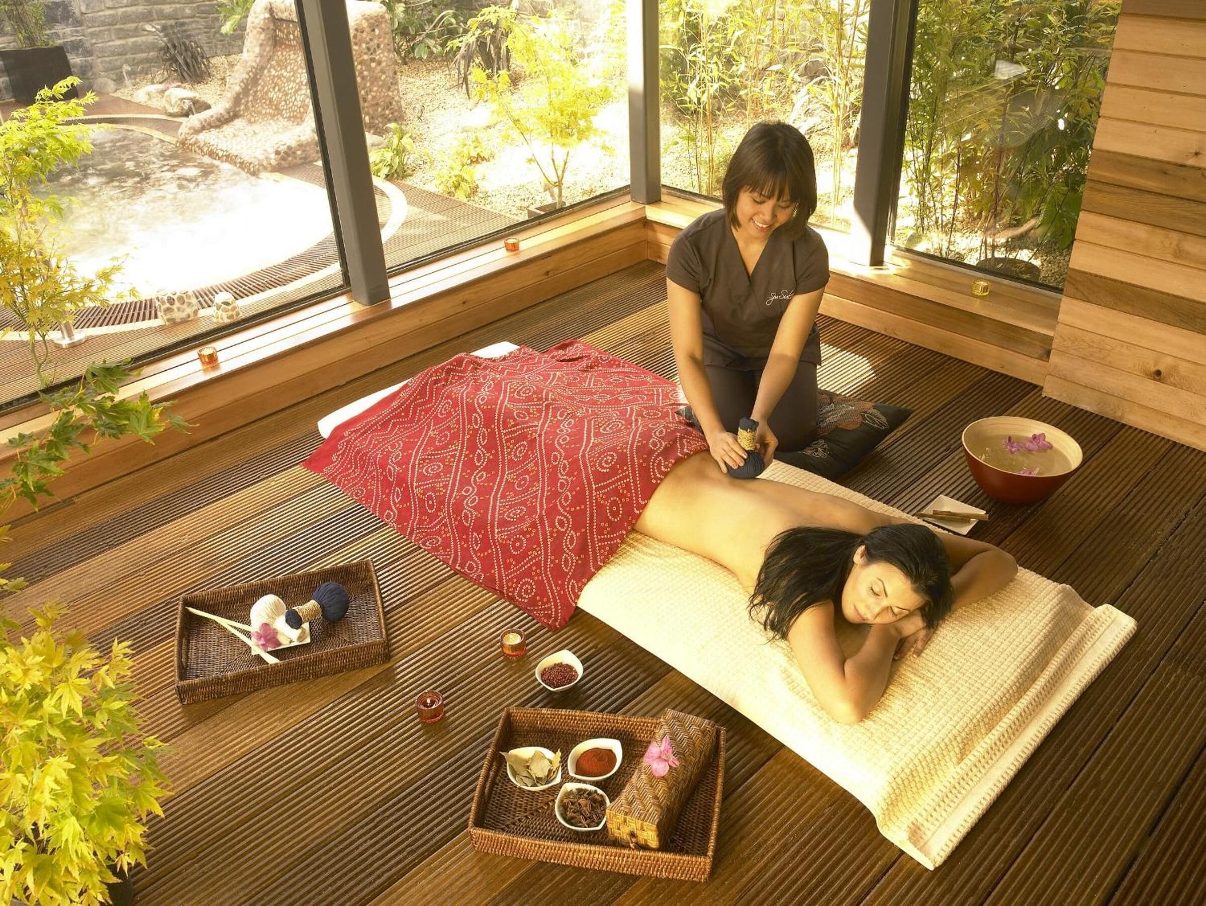 A spa room with a wooden floor, a red patterned mat, a massage table, and a window with a view of greenery.