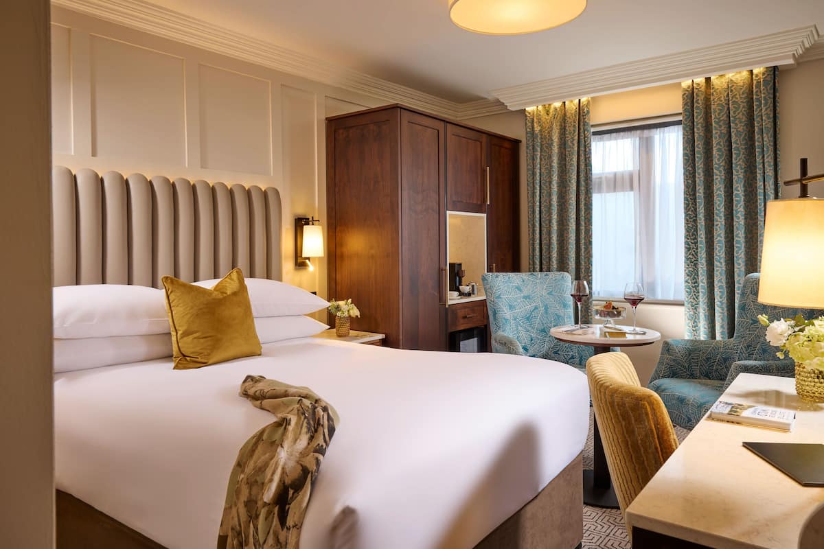 Ein Hotelzimmer mit einem großen Bett, einem Holzschrank, einem kleinen runden Tisch mit Lampe, einem Sessel und einem Fenster mit Vorhängen.