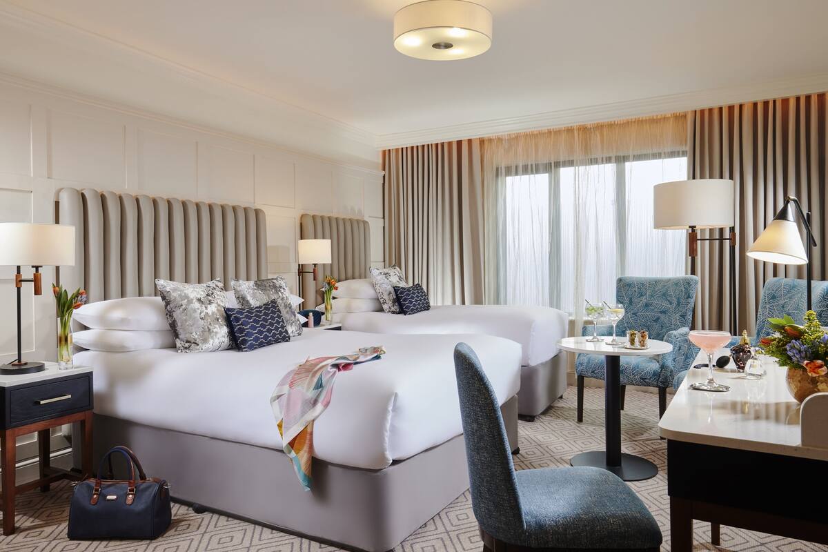 Ein Hotelzimmer mit einem großen Bett, zwei Nachttischen mit Lampen, einem Sessel, einem kleinen Tisch mit Getränken und Blick auf die Stadt durch große Fenster.