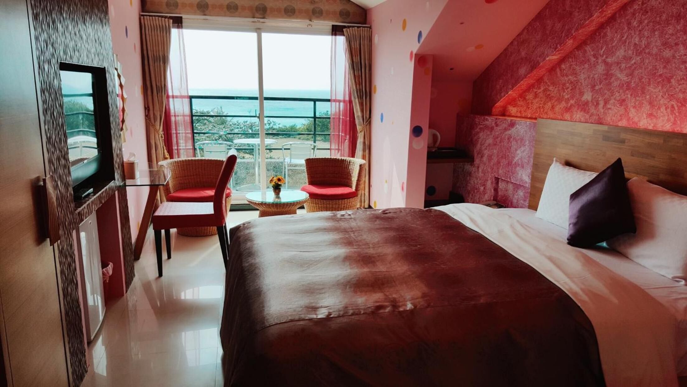 Habitación de hotel con una cama grande, televisión, dos sillas, una mesita y un balcón con vistas.